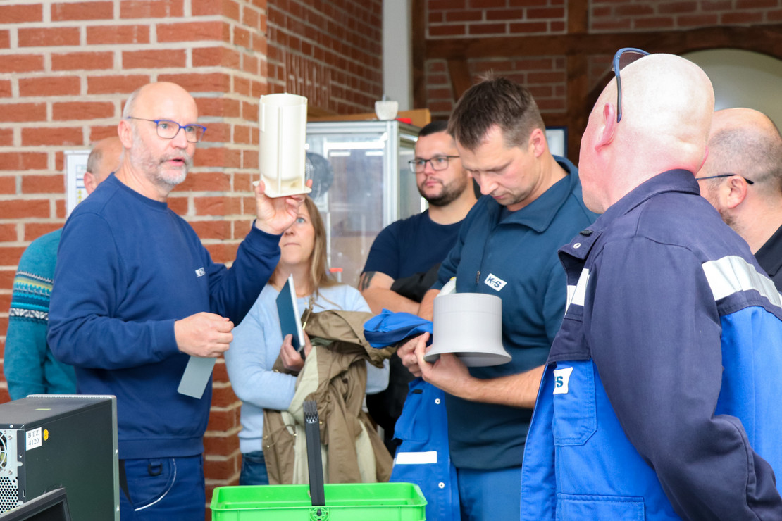 Mehrere Personen in blauer Arbeitskleidung stehen in einer Werkstatt und unterhalten sich. Eine Person zeigt einer Gruppe einen 3D-Druck.
