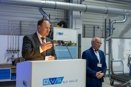 Ein Man im schwarzen Anzug mit gestreifter Krawatte spricht an einem Rednerpult mit DVS SLV Halle-Logo, im Hintergrund befindet sich ein Roll-up mit Leistungsübersicht der SLV Halle GmbH sowie technische Anlagen in einer Werkhalle. Daneben steht ein Mann mit blauem Anzug.