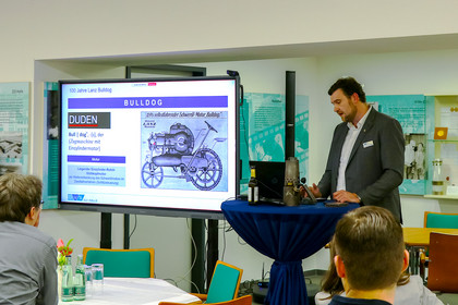 Ein Mann im Anzug spricht an einem Stehtisch mit historischem Exponat über den Lanz Bulldog. Hinter ihm ist eine Präsentationsfolie mit einer technischen Zeichnung und Definition des Begriffs „Bulldog“ zu sehen.