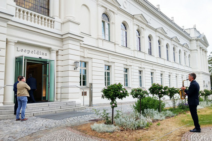 Ein Musiker spielt Saxophon vor dem Haupteingang der Leopoldina in Halle (Saale), während mehrere Personen das Gebäude betreten.