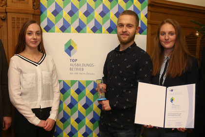 Zwei Frauen und ein Mann stehen vor einem Banner, der Mann hält eine Urkunde in den Händen, alle drei lächeln. Auf den Banner steht: "Top Ausbildungsbetrieb der IHK Halle-Dessau"