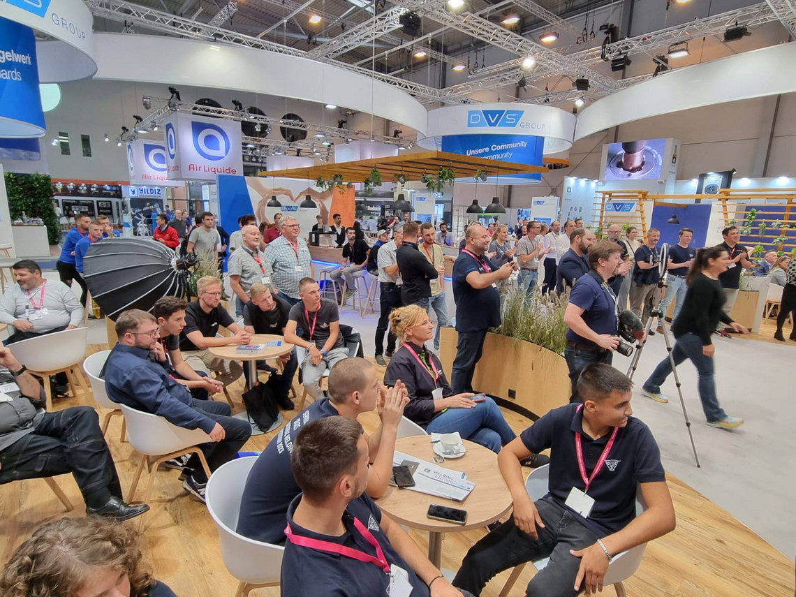 Zahlreiche Männer und Frauen sitzen und stehen auf einem Messestand der DVS Group, verfolgen aufmerksam eine Präsentation oder Vorführung; im Vordergrund befinden sich Sitzgruppen mit Holztischen, im Hintergrund Kameratechnik und Firmenbanner.