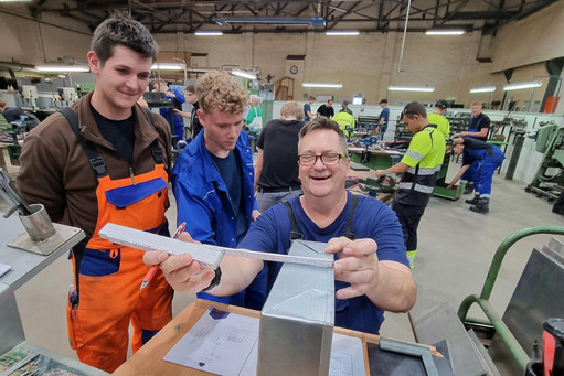 Zwei Azubis stehen an einem Arbeitsplatz in einer Werkhalle und ihr Ausbilder vermisst begeistert ein Metallbauteil. Im Hintergrund befinden sich mehrere weitere Personen sowie Arbeitsplätze und technische Geräte.