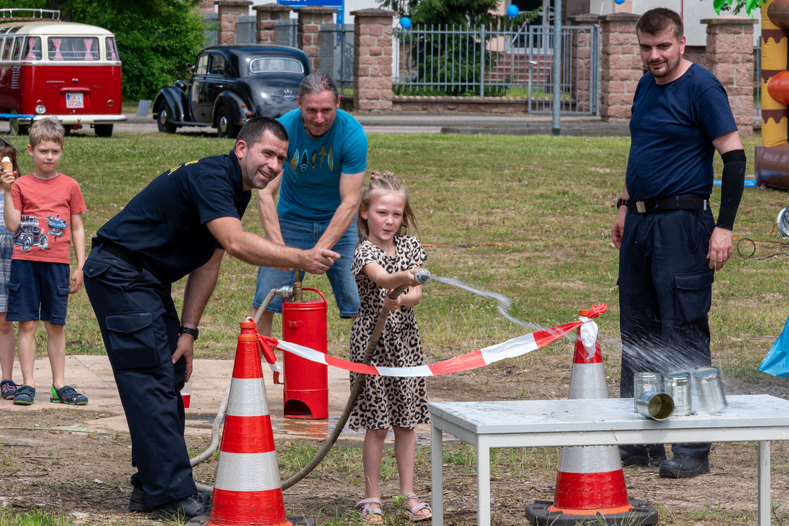 Drei Erwachsene und zwei Kinder stehen an einem Aktionsstand im Freien und beobachten, wie ein Mädchen unter der Anleitung eines Feuerwehrmannes konzentriert mit einem Wasserschlauch auf eine Pyramide aus Dosen zielt.