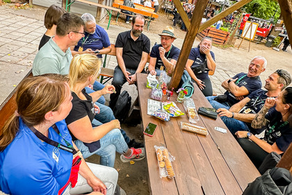 Eine größere Gruppe von Personen sitzt lachend im Freien an einem Holztisch. Auf dem Tisch liegen Getränkeflaschen, Verpackungen und weitere Gegenstände, im Hintergrund sind Bäume und Sitzbereiche zu sehen.