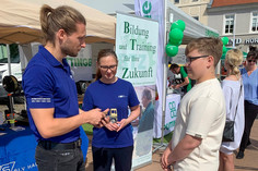 Ein Mann und eine Frau im blauen SLV-Halle-Shirt sprechen mit einem Jugendlichen vor einem Messestand im Freien, im Hintergrund ist ein Banner mit der Aufschrift „Bildung und Training für Ihre Zukunft“ zu sehen.