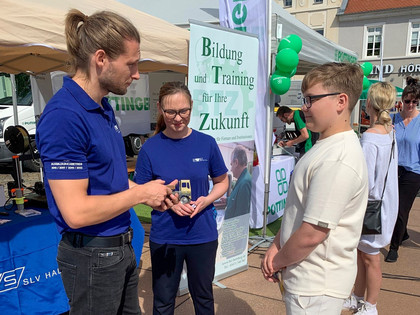 Ein Mann und eine Frau im blauen SLV-Halle-Shirt sprechen mit einem Jugendlichen vor einem Messestand im Freien, im Hintergrund ist ein Banner mit der Aufschrift „Bildung und Training für Ihre Zukunft“ zu sehen.