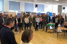 Besucherinnen und Besucher stehen dicht gedrängt in einem Messebereich mit Informationsständen, einige tragen Uniformen, andere Namensschilder, vorne ist ein Tisch mit Auslagen und der Standnummer 18 zu sehen.