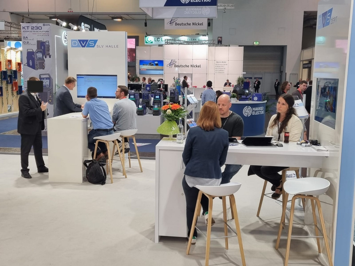 Besuchende nterhalten sich an mehreren weißen Stehtischen auf dem Messestand der DVS SLV Halle, im Hintergrund sind weitere Aussteller und Schweißtechnik-Exponate zu sehen.