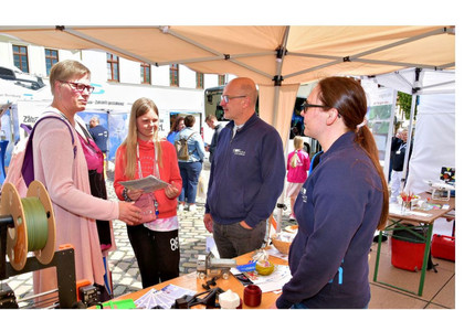 Fünf Personen stehen unter einem Pavillon auf einer Messe im Gespräch. Zwei Frauen und ein Mädchen informieren sich an einem Stand mit 3D-Druckmaterialien, während zwei Standbetreuende in blauer Kleidung erklären. Im Vordergrund sind Druckteile und Filamentrollen zu sehen.