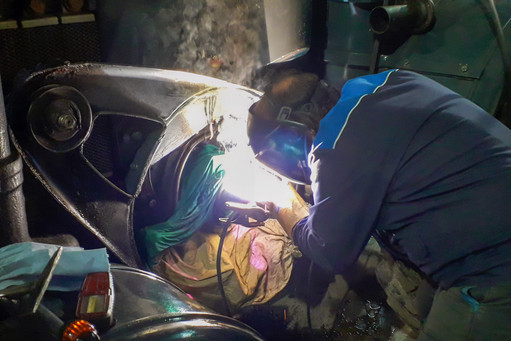 Eine Person in Schutzkleidung führt eine Schweißarbeit an einem großen Metallbauteil durch; grelles Licht und Funken zeigen den aktiven Schweißvorgang, im Hintergrund sind Maschinenteile und Werkzeuge sichtbar.