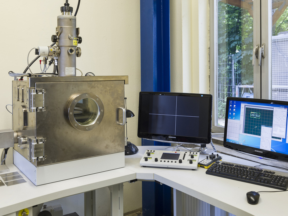 Metallene Vakuumkammer mit Sichtfenster steht auf einem Labortisch neben zwei Monitoren, einem Steuerpult und einer Tastatur; durch das Fenster im Hintergrund ist Grünfläche sichtbar.