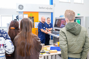 Mehrere Jugendliche stehen an einem Arbeitstisch mit technischen Bauteilen und Messgeräten in einer Werkhalle. Zwei Männer in blauer Arbeitskleidung erklären einen Prüfaufbau, während die anderen Personen den Tisch und einen Bildschirm betrachten.