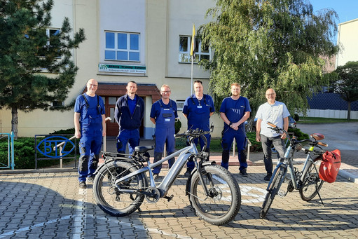 Fünf Männer in blauer Arbeitskleidung und ein Mann in Zivil stehen auf einem Firmenparkplatz vor einem Gebäude, zwei E-Bikes mit Gepäcktaschen sind im Vordergrund abgestellt.