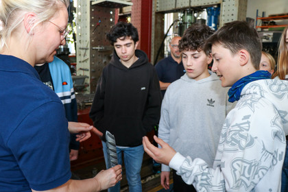 Drei Jungen stehen in einer Werkhalle um einen Arbeitstisch und betrachten gemeinsam ein zerbrochenes Stück Betonstahl, den eine Frau in der Hand hält. Im Hintergrund sind Maschinen und technische Anlagen sichtbar.
