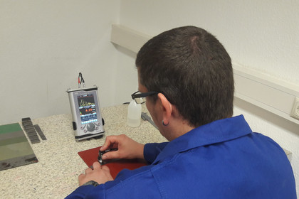 Ein Mann in blauer Arbeitskleidung führt an einem Tisch eine Prüfung mit einem tragbaren Ultraschallprüfgerät durch. Er hält eine kleine Sonde auf ein flaches Werkstück, auf dem Bildschirm sind Messkurven sichtbar.
