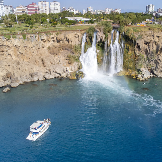 Boot auf dem Meer vor den Düden-Wasserfällen bei Antalya, die von einer Klippe direkt ins Mittelmeer stürzen, im Hintergrund Stadtgebäude.