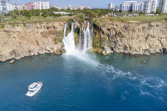 Boot auf dem Meer vor den Düden-Wasserfällen bei Antalya, die von einer Klippe direkt ins Mittelmeer stürzen, im Hintergrund Stadtgebäude.