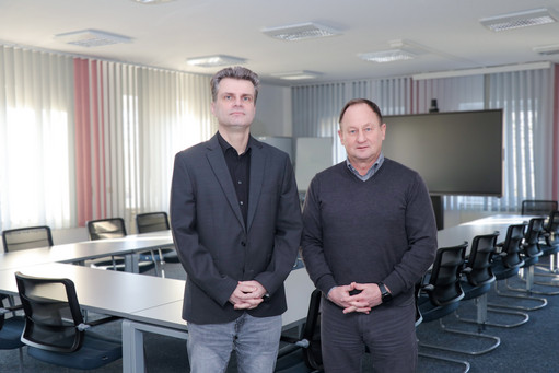 Zwei Männer stehen in einem modernen, hellen Konferenzraum mit U-förmig angeordneten Tischen und schwarzen Bürostühlen; im Hintergrund ein großer Bildschirm und vertikale Lamellenvorhänge.