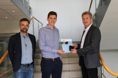 Drei Männer stehen auf einer Treppe in einem modernen Gebäude, der mittlere erhält eine Urkunde und einen kleinen Karton von einem der anderen; alle tragen Businesskleidung.