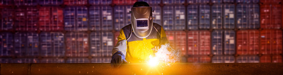Schweißer mit Schutzhelm arbeitet vor einer Wand aus bunten Containern, heller Funkenflug leuchtet die Szene aus.