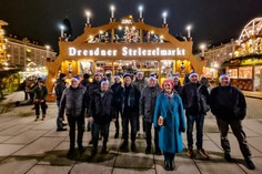 Eine Gruppe von Mitarbeitenden der SLV Halle GmbH und der SLV Hannover steht vor dem beleuchteten Eingang des Dresdner Striezelmarkts. Die Teilnehmenden tragen festliche Mützen und genießen die weihnachtliche Atmosphäre.