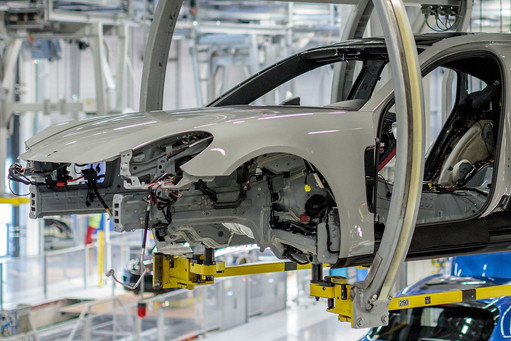 Unlackierte Karosserie eines Fahrzeugs hängt in automatisierter Montagelinie, sichtbare Kabel und Montagetechnik in moderner Produktionshalle