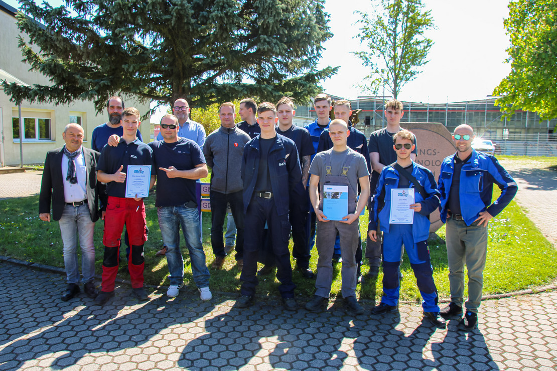 Gruppenfoto mit 17 Personen im Außenbereich eines Ausbildungszentrums; vorn stehen mehrere junge Männer in Arbeitskleidung mit Urkunden, dahinter Ausbilder und weitere Teilnehmende, im Hintergrund ein großer Nadelbaum und ein Gebäudekomplex.