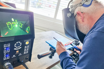 Ein Mann arbeitet an einem Elektronikarbeitsplatz und hält ein kleines Bauteil mit einer Pinzette, während er mit einem Lötkolben daran arbeitet. Vor ihm steht ein Monitor, der eine vergrößerte Ansicht der Lötstelle zeigt, daneben befinden sich Geräte und Werkzeuge auf dem Tisch.