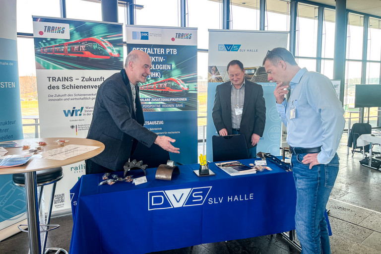 Zwei Männer stehen an einem Informationsstand der SLV Halle GmbH und betrachten metallische Bauteile auf einem Tisch mit blauem DVS SLV Halle Tischtuch. Hinter ihnen stehen Roll-ups zu TRAINS und zum Lehrgang Gestalter PTB Perspektivtechnologien IHK. Ein weiterer Mann steht im Hintergrund vor einem Roll-up der SLV Halle. Große Fensterfront eines Veranstaltungsgebäudes im Hintergrund.