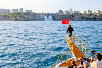 Eine Person steht am Ende eines gelben Schiffsanlegers mit einer türkische Flagge über dem Wasser. Im Hintergrund sind ein Wasserfall, bewaldete Uferbereiche und mehrere Hochhäuser zu sehen, im Vordergrund sitzen weitere Personen auf einem Boot.