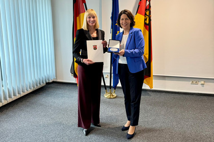 Zwei Frauen stehen in einem Veranstaltungsraum, eine überreicht der anderen eine Urkunde, im Hintergrund sind Fahnen von Deutschland, der EU und Rheinland-Pfalz vor einer hellen Wand zu sehen.