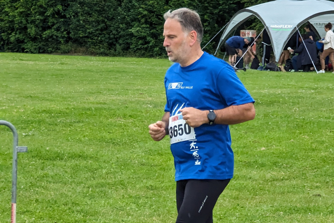 Ein Mann im blauen SLV Halle GmbH-T-Shirt mit Startnummer 3650 läuft über eine Wiese bei einer Laufveranstaltung. Im Hintergrund stehen Menschen vor einem weißen Zelt.
