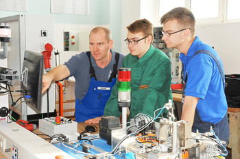 Ein Ausbilder zeigt zwei Auszubildenden an einem Bildschirm technische Abläufe einer Steuerungsanlage in einer Werkstattumgebung.
