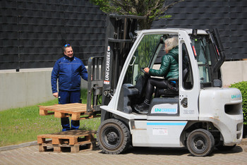 Eine Frau steuert einen Gabelstapler mit angehobener Holzpalette, während ein Mann in blauer Arbeitskleidung daneben steht und den Vorgang beobachtet © SLV Halle GmbH 2025