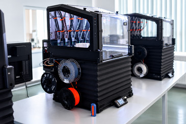 Zwei 3D-Drucker mit transparenten Gehäusen und seitlich angebrachten Filamentrollen stehen auf einem Arbeitstisch  