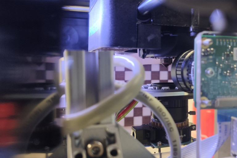 Nahaufnahme eines technischen Prüfaufbaus mit mehreren Metallbauteilen, Schläuchen und einer Kamera. Rechts befindet sich ein elektronisches Gerät mit Display.