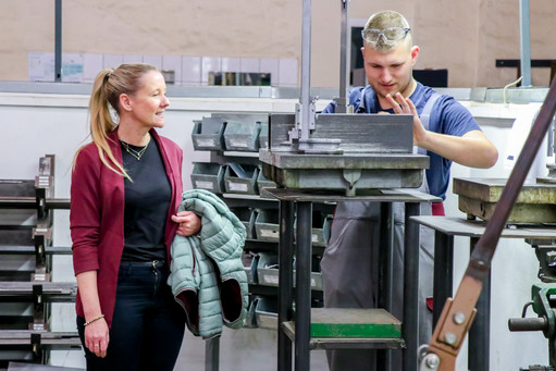 Eine Frau und ein Mann stehen in einer Werkstatt an einer Metallmaschine. Der Mann erklärt etwas, während die Frau aufmerksam zuhört.