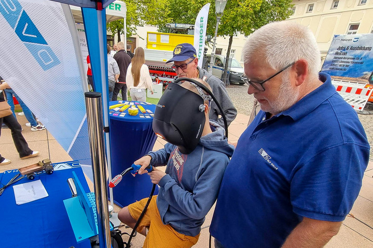 Ein Mann mit grauen Haaren und Brille steht an einem Informationsstand und betrachtet gemeinsam mit einer weiteren Person ausgestellte Gegenstände auf einem blau gedeckten Tisch. Im Hintergrund sind mehrere Personen, Banner und weitere Stände im Außenbereich zu sehen.