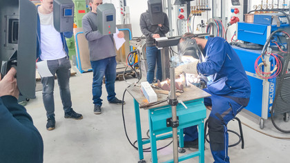 Ein Ausbilder demonstriert eine Schweißtechnik an einem Werkstück auf einem Tisch, während fünf Teilnehmende mit Schweißschilden und Unterlagen im Halbkreis zuschauen. Im Hintergrund stehen technische Geräte und Gasflaschen.