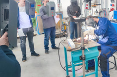 Ein Ausbilder demonstriert eine Schweißtechnik an einem Werkstück auf einem Tisch, während fünf Teilnehmende mit Schweißschilden und Unterlagen im Halbkreis zuschauen. Im Hintergrund stehen technische Geräte und Gasflaschen.
