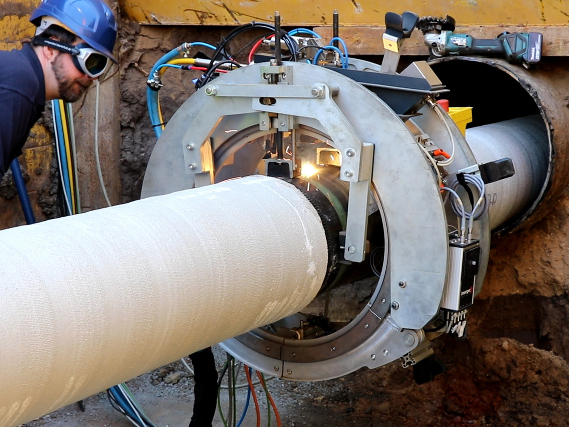Ein Techniker mit Schutzbrille und Helm überwacht ein orbitales Schweißsystem, das zwei große Rohre in einem Erdloch verbindet; das Gerät ist rund um die Rohrverbindung montiert, Kabel und Sensoren führen zum Steuergerät.