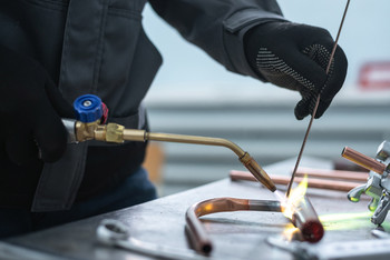 Person mit schwarzen Handschuhen führt eine Lötflamme an ein Kupferrohr, während mit einem Zusatzdraht gearbeitet wird.