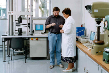 Zwei Personen in Arbeitskleidung stehen in einer Werkhalle an einer Reihe stationärer Maschinen und betrachten gemeinsam ein Werkstück an einem Arbeitsplatz mit Werkzeugen.