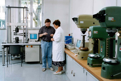 Zwei Personen in Arbeitskleidung stehen in einer Werkhalle an einer Reihe stationärer Maschinen und betrachten gemeinsam ein Werkstück an einem Arbeitsplatz mit Werkzeugen.