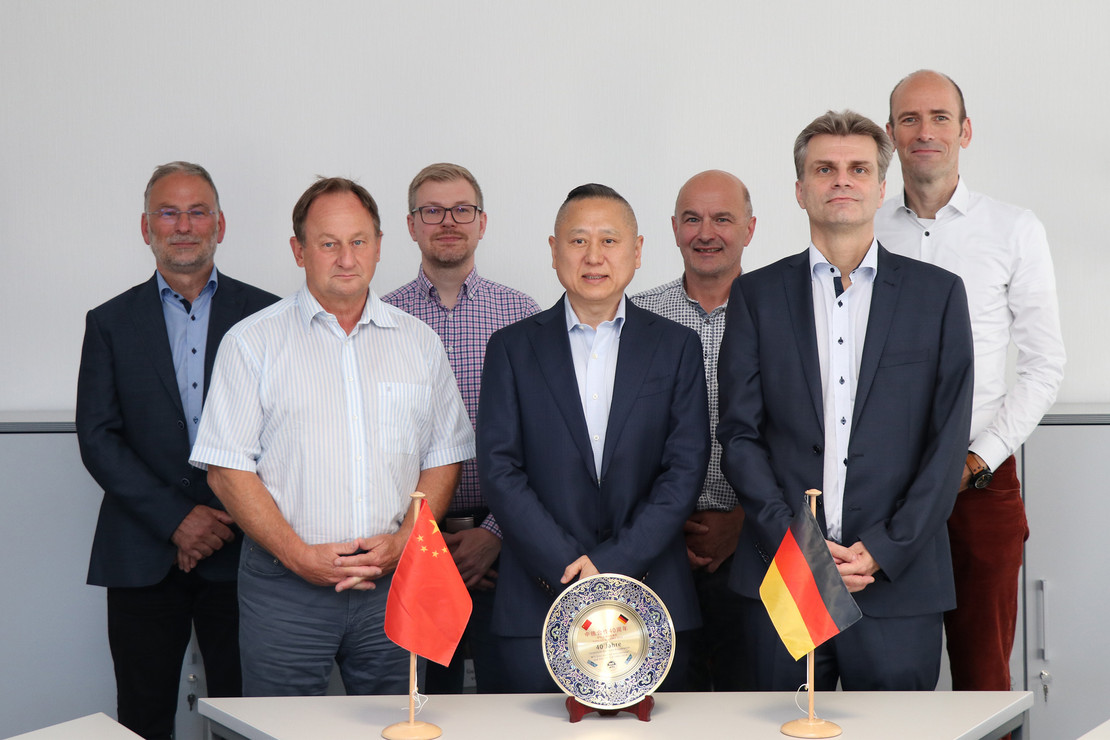 Gruppenfoto vor dem Gastgeschenk der WTI Harbin, China. Der dekorative Teller steht auf einem Tisch mit kleinen Flaggen von China und Deutschland.