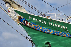 Nahaufnahme des grünen Schiffsbugs der „Rickmer Rickmers“ mit bunter Verzierung, Namenszug und weißer Galionsfigur vor blauem Himmel