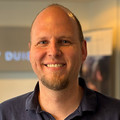 Ein Mann mit Glatze und kurzem Bart lächelt in die Kamera. Er trägt ein dunkelblaues Poloshirt, der Hintergrund ist unscharf und zeigt einen Innenraum mit verschwommenen Elementen.