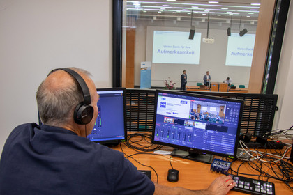 Ein Techniker mit Kopfhörer steuert in einem Regieraum über zwei Monitore und ein Mischpult die Videoübertragung einer Konferenz, die durch ein Fenster in den Saal mit Präsentationseinblendung sichtbar ist.
