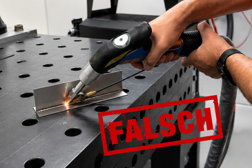 Eine Person führt Schweißarbeiten an einem Metallbauteil auf einer Werkbank aus. Im Vordergrund ist der rote Schriftzug „FALSCH“ zu sehen, die Person trägt keine erkennbaren Schutzhandschuhe.
